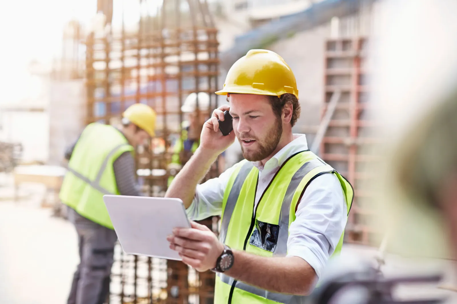 Contractors on a construction site. One is on the phone while looking at a tablet.
