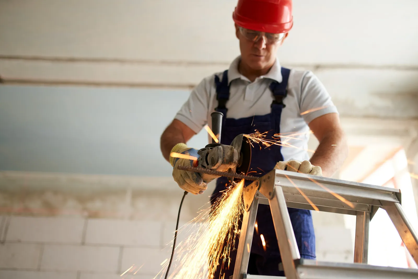 Professional construction worker hands in work gloves using angle grinder to cut metal rod at building site. Close up shot of contractor cutting iron stick on a ladder with glowing sparks.