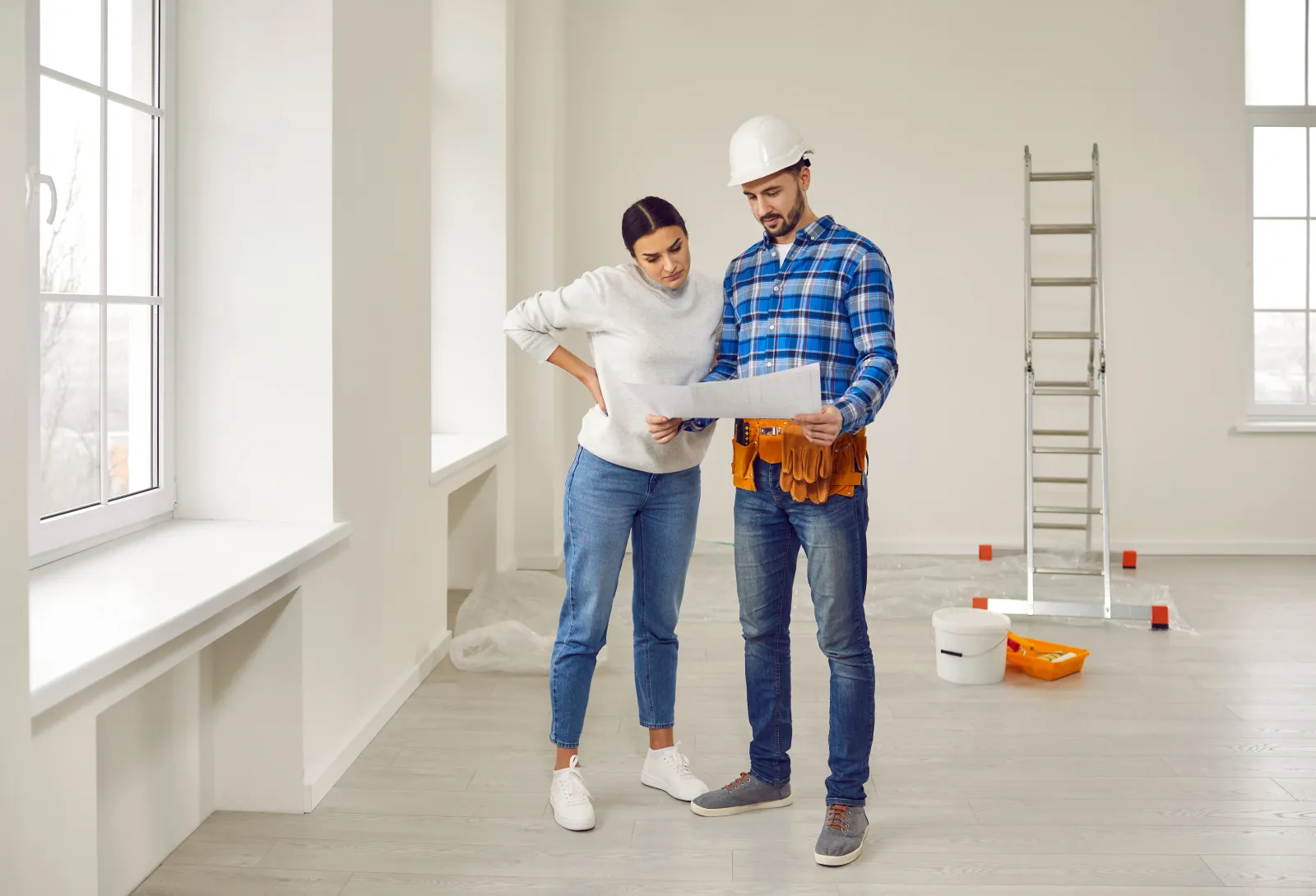 Finishing painting decorating work foreman builder supervisor in hardhat showing young woman homeowner renovation plan standing in empty house or apartment with ladder and plaster bucket in background